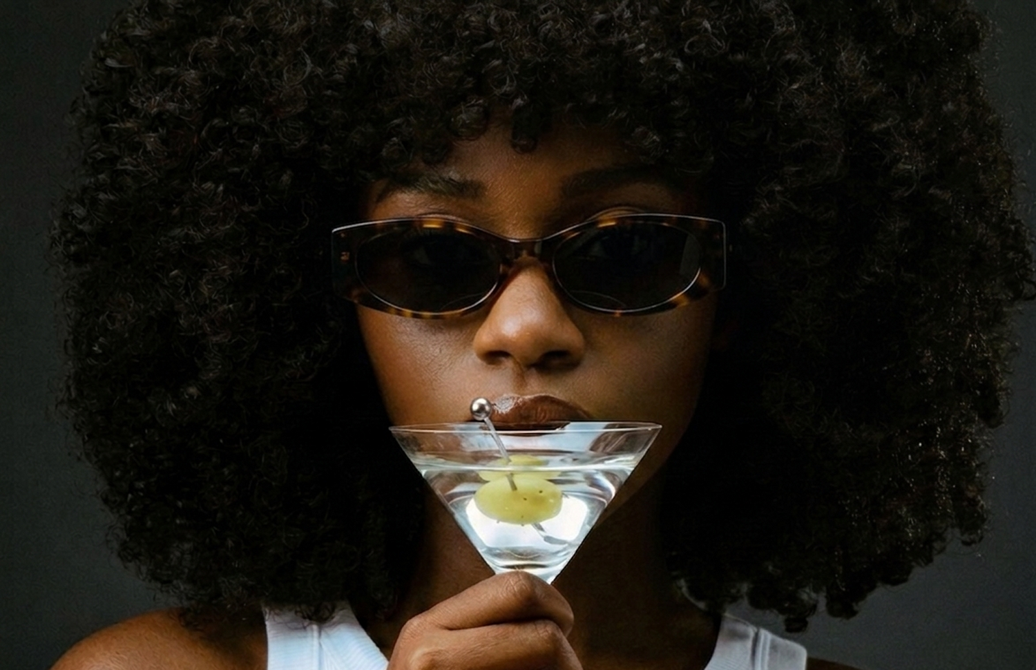 Person in sunglasses holding a martini glass with a olive against a dark background